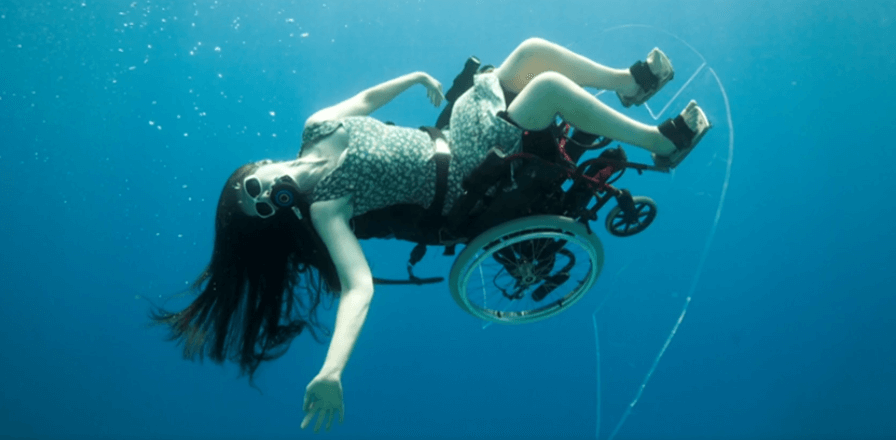 A woman in a floral dress and scuba mask, strapped into a wheelchair, poses underwater. She floats parallel to the surface of the water, arms and hair outstretched.