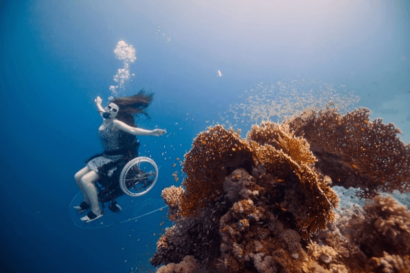 A woman glides underwater in her wheelchair and scuba mask. She sits upright and turns around a mass of orange coral, arms outstretched with a trail of bubbles behind and above her.