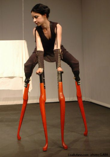 A dancer perches with four limbs each affixed to a tall orange prosthetic stilt.