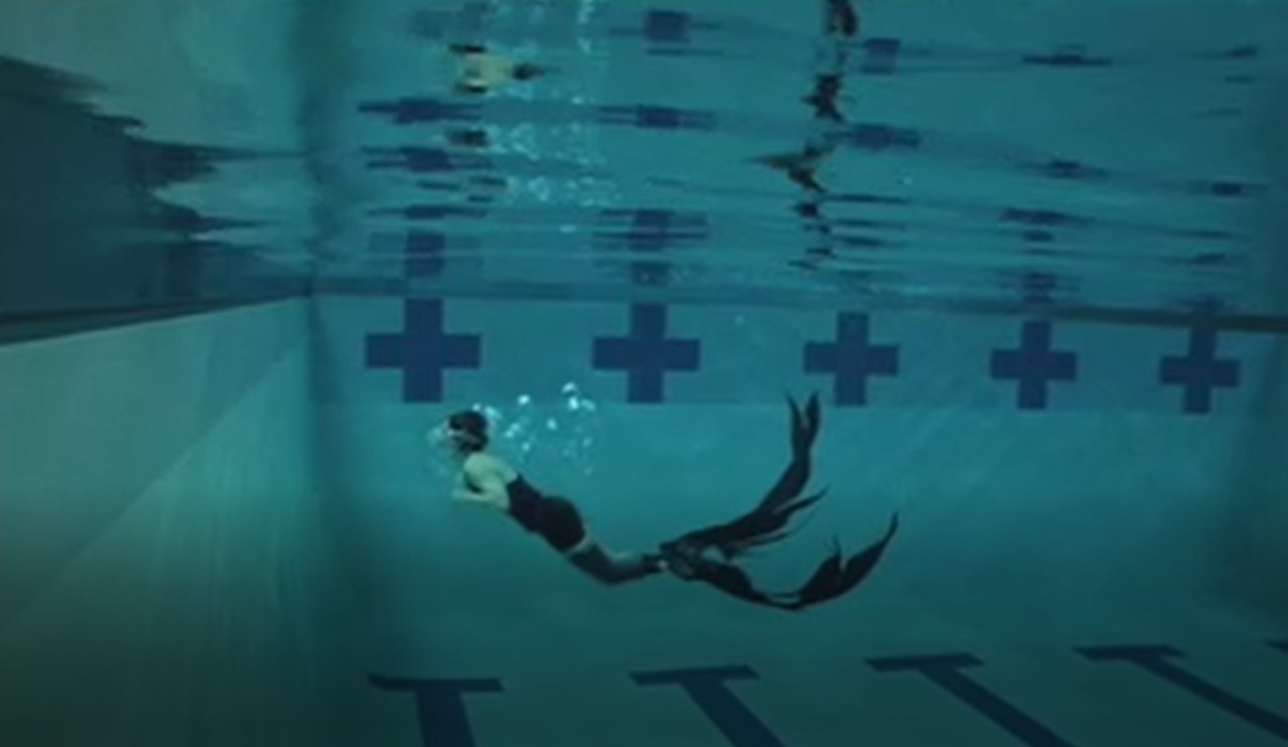 A woman swims underwater toward the surface of a pool with a feathery mono-fin that billows through the water.