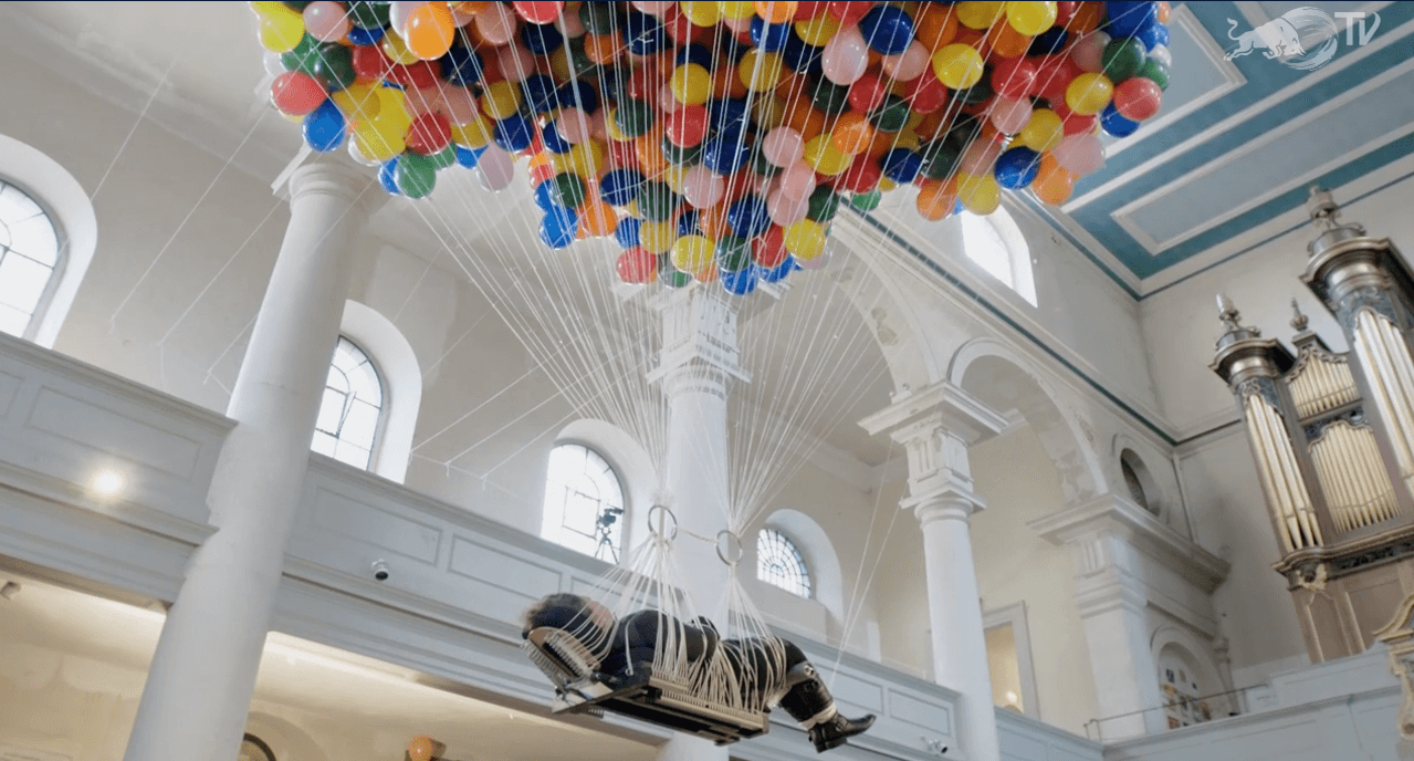 A woman lies on a flat board to which hundreds of colorful party balloons are tied. She floats through the air, near the ceiling of a white-walled church.