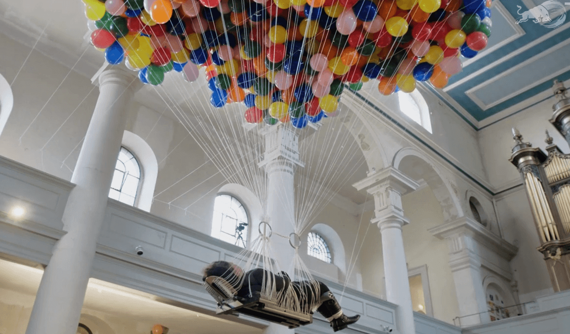 A woman lies on a flat board to which hundreds of colorful party balloons are tied. She floats through the air, near the ceiling of a white-walled church.