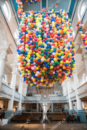 A block of 20000 rainbow balloons float in an open church, with strings leading toward a small figure of a woman laying on a flat board.