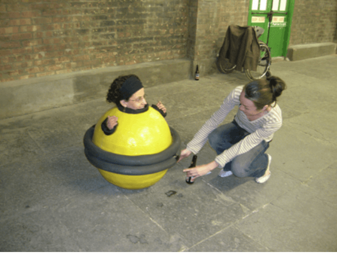 A woman rests within a small yellow spherical performance-object. Only her head and hands come outside of the spherical body. A second woman crouches next to her, arms reaching out towards the sphere as if to push it.