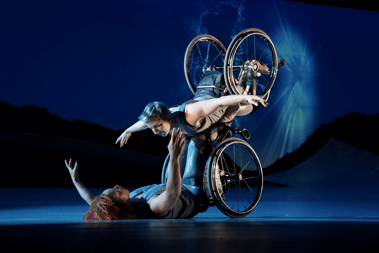 A dancer balances in the air, resting on the footplate of another dancer's wheelchair. Both face each other with arms outstretched against the backdrop of a starry night.
