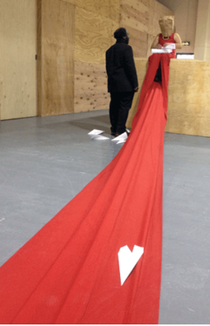 A woman sits with a paper bag over her head, wearing a long red dress, folding and throwing paper airplanes which sit scattered around her. A man dressed in black stands close by her, looking up to her covered face.