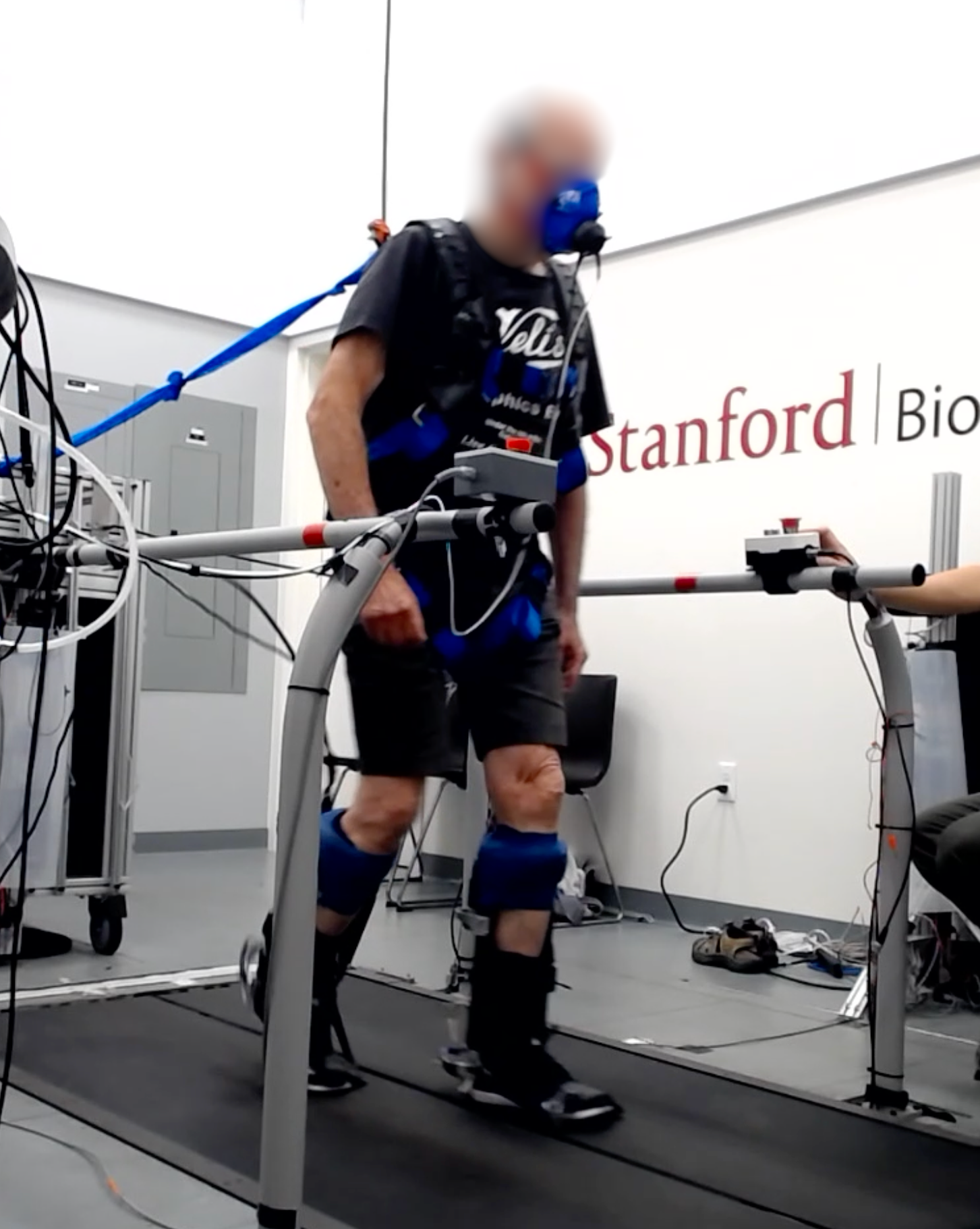 A blurred-faced man walks on a treadmill in a laboratory setting, wearing ankle exoskeletons on both feet, a safety harness, and respiratory mask. 